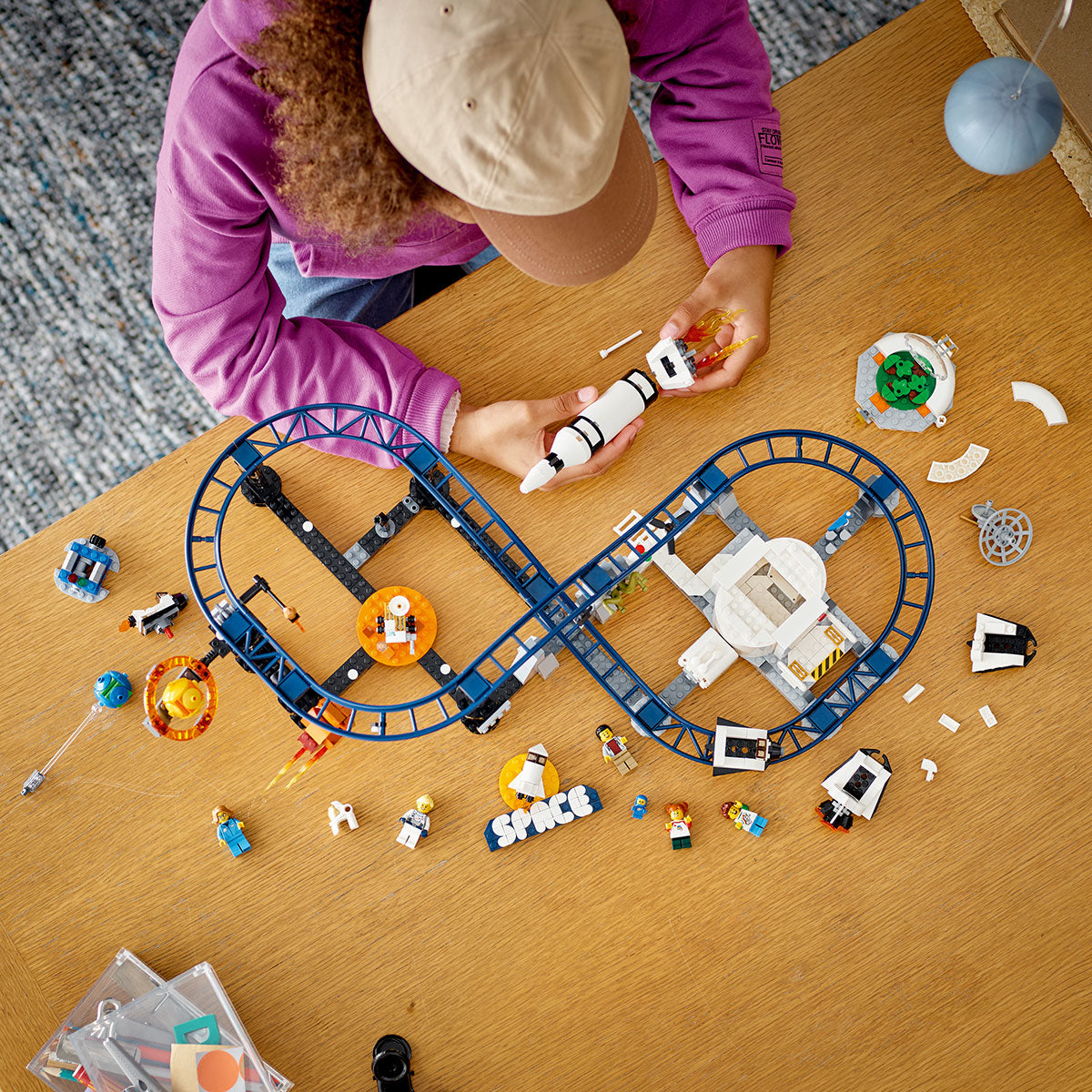 A child in a pink long-sleeve shirt and beige cap builds the LEGO® Space Roller Coaster by LEGO, featuring blue tracks, miniature astronauts, rockets, and space accessories—perfect for young space fans—on a wooden table.
