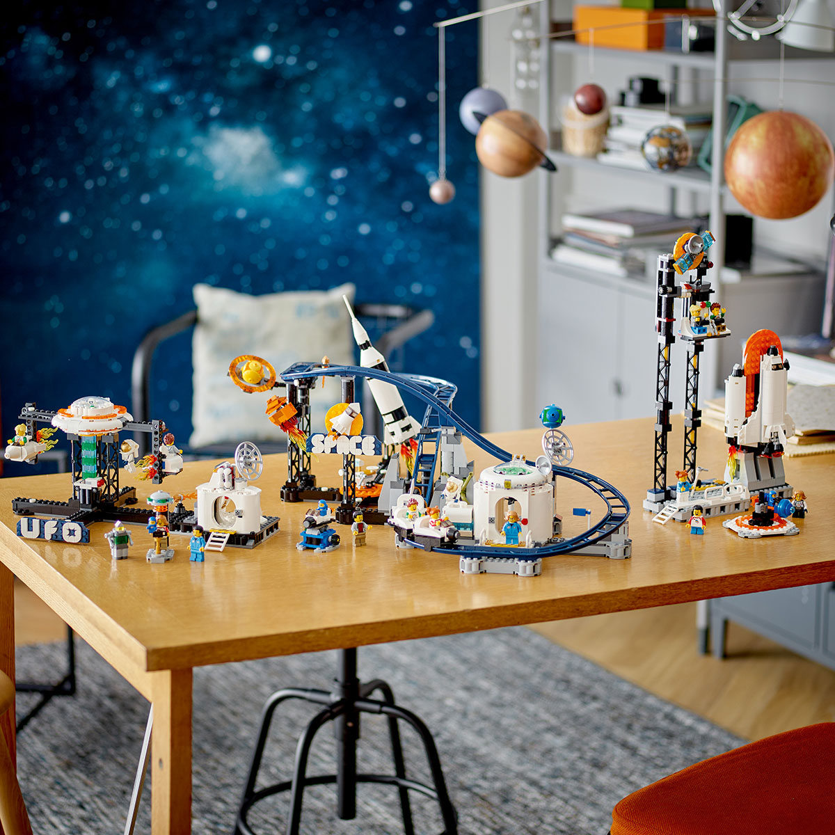 A table displays various LEGO space sets—rockets, astronauts, planets, vehicles, and the LEGO® Space Roller Coaster by LEGO—while a space mural and hanging solar system model set a cozy scene for fans.