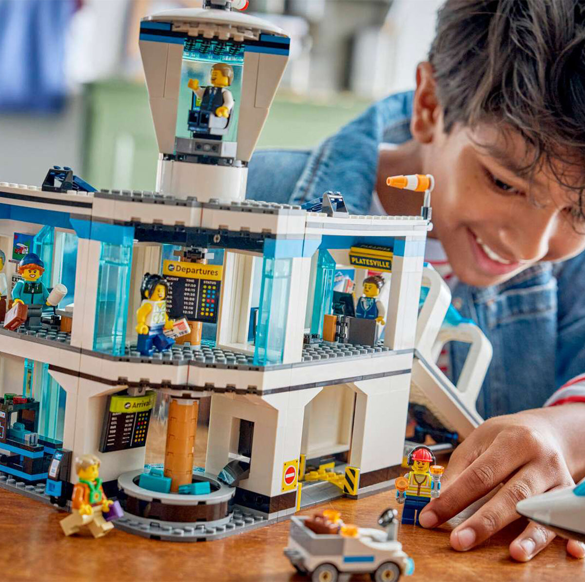 A smiling child engages in imaginative play with the BOEING LEGO® City Passenger Airplane set, featuring minifigures, a control tower, and various airport elements on a table.