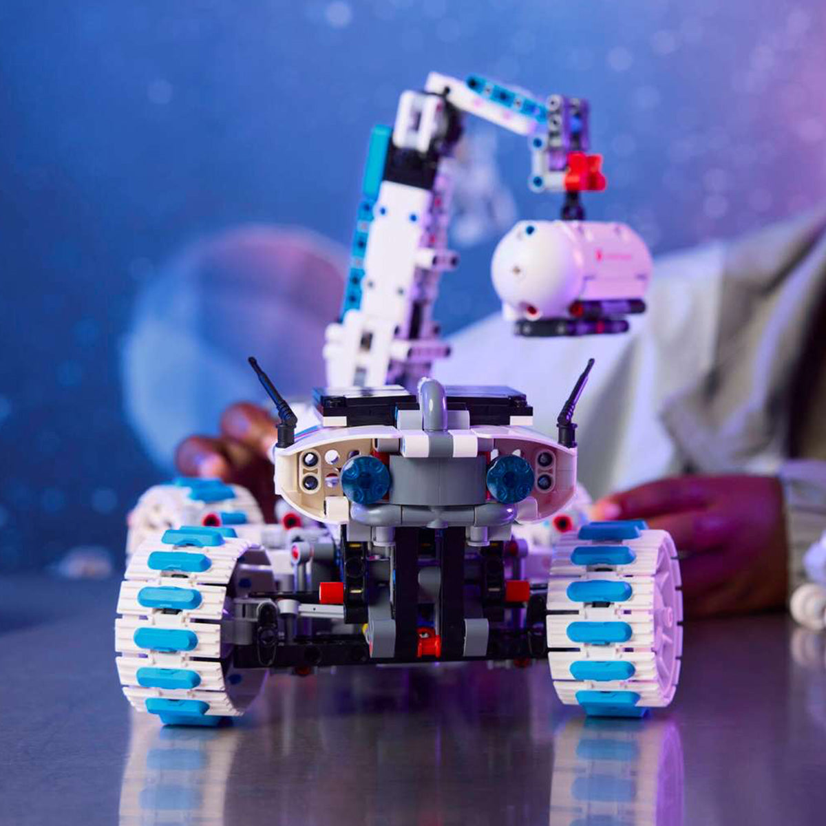 A close-up of the BOEING LEGO® Technic Lunar Outpost Moon Rover, featuring large white and blue wheels and a mechanical arm, being operated by a person in a lab coat against a space-themed background.