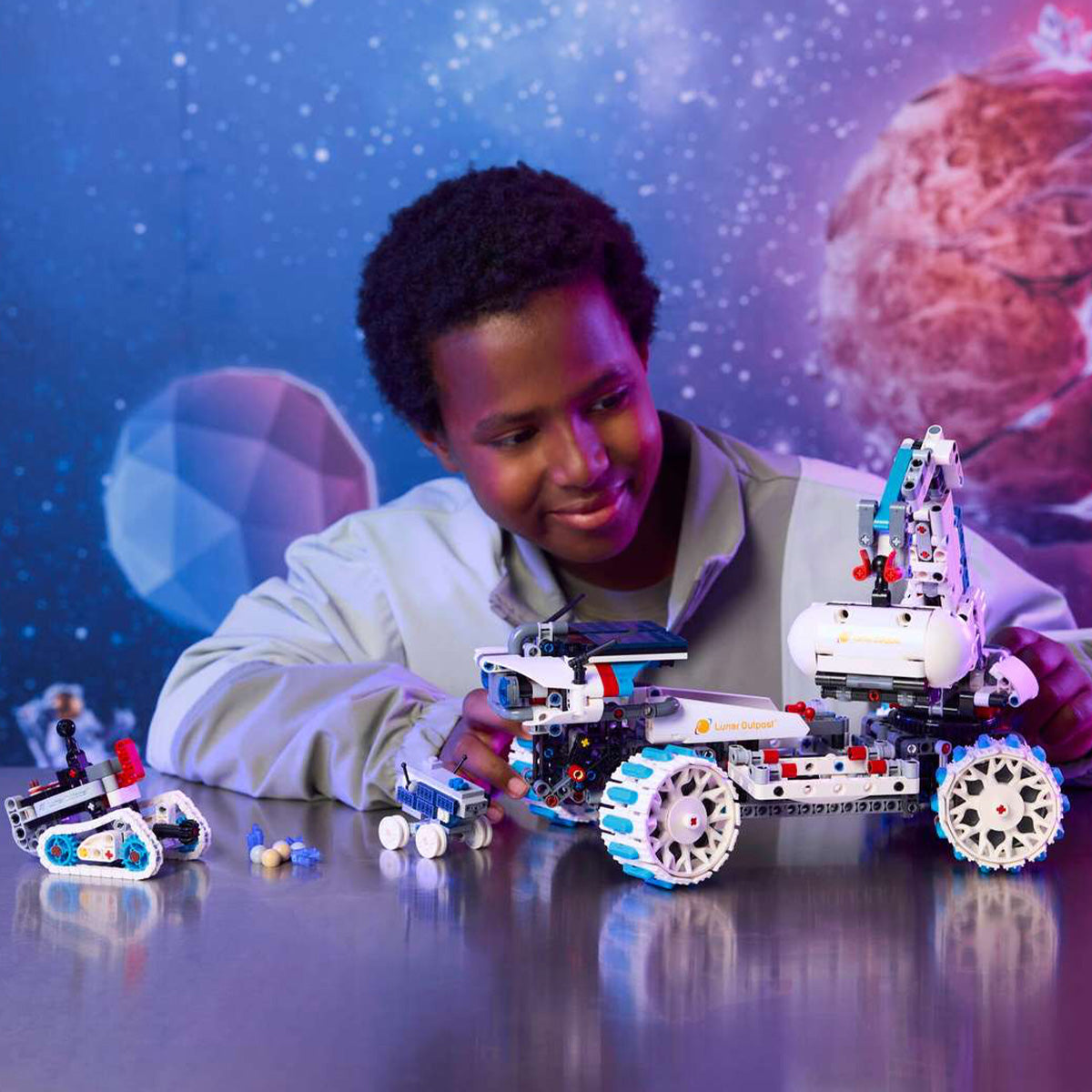 A smiling child assembles a BOEING LEGO® Technic Lunar Outpost Moon Rover on a table, with two smaller Moon Rover models and minifigures nearby. The background features a vibrant, space-themed mural with planets and stars, capturing the excitement of building this detailed lunar vehicle construction set.