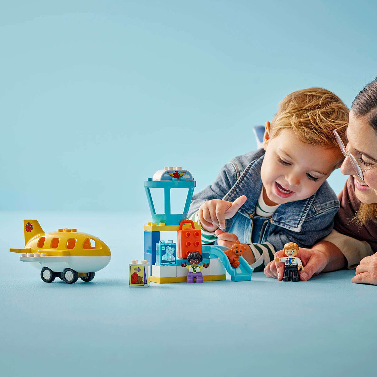 A smiling child and adult engage in imaginative play with the LEGO® DUPLO® First Time At The Airport set by BOEING, featuring a toy airplane, control tower, and figurines on a blue surface against a light blue background—perfect for little aviators exploring their very first airport adventure.