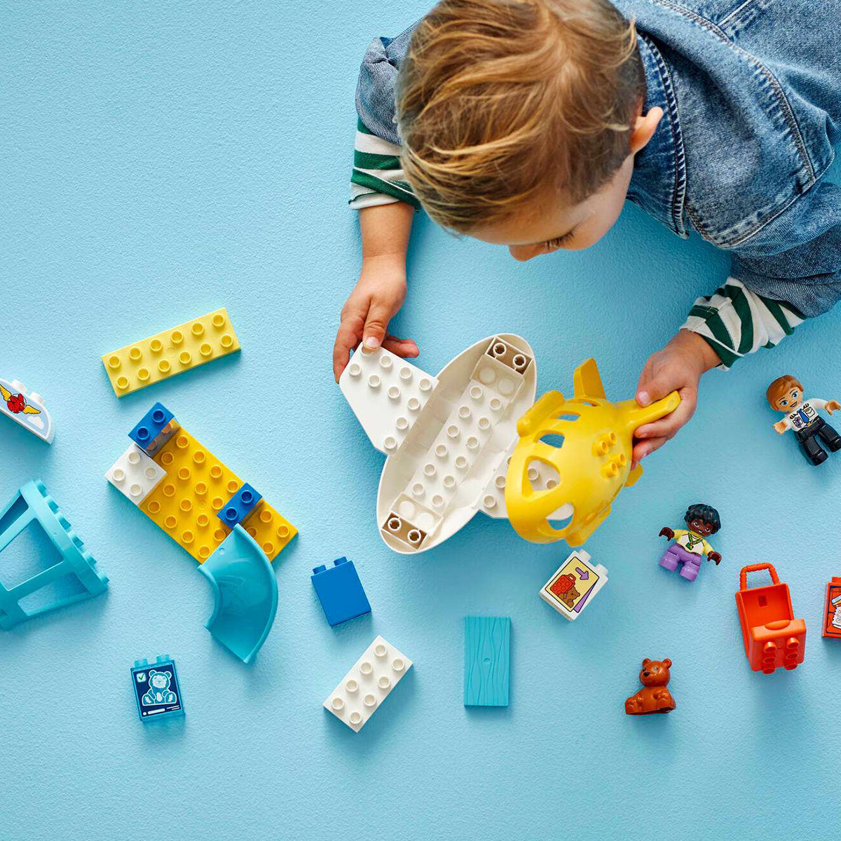 A child plays with the colorful LEGO® DUPLO® First Time At The Airport set by BOEING, featuring airport building blocks, a toy airplane, and small figurines on a blue surface—perfect for recreating their first trip to the airport with this toddler building set.