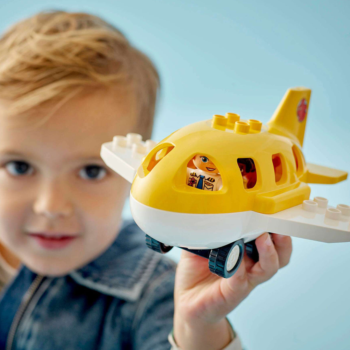 A young child with light brown hair holds up the BOEING LEGO® DUPLO® First Time At The Airport set, featuring a bright yellow toddler airplane and a small figure inside, smiling at the camera against a light blue background.