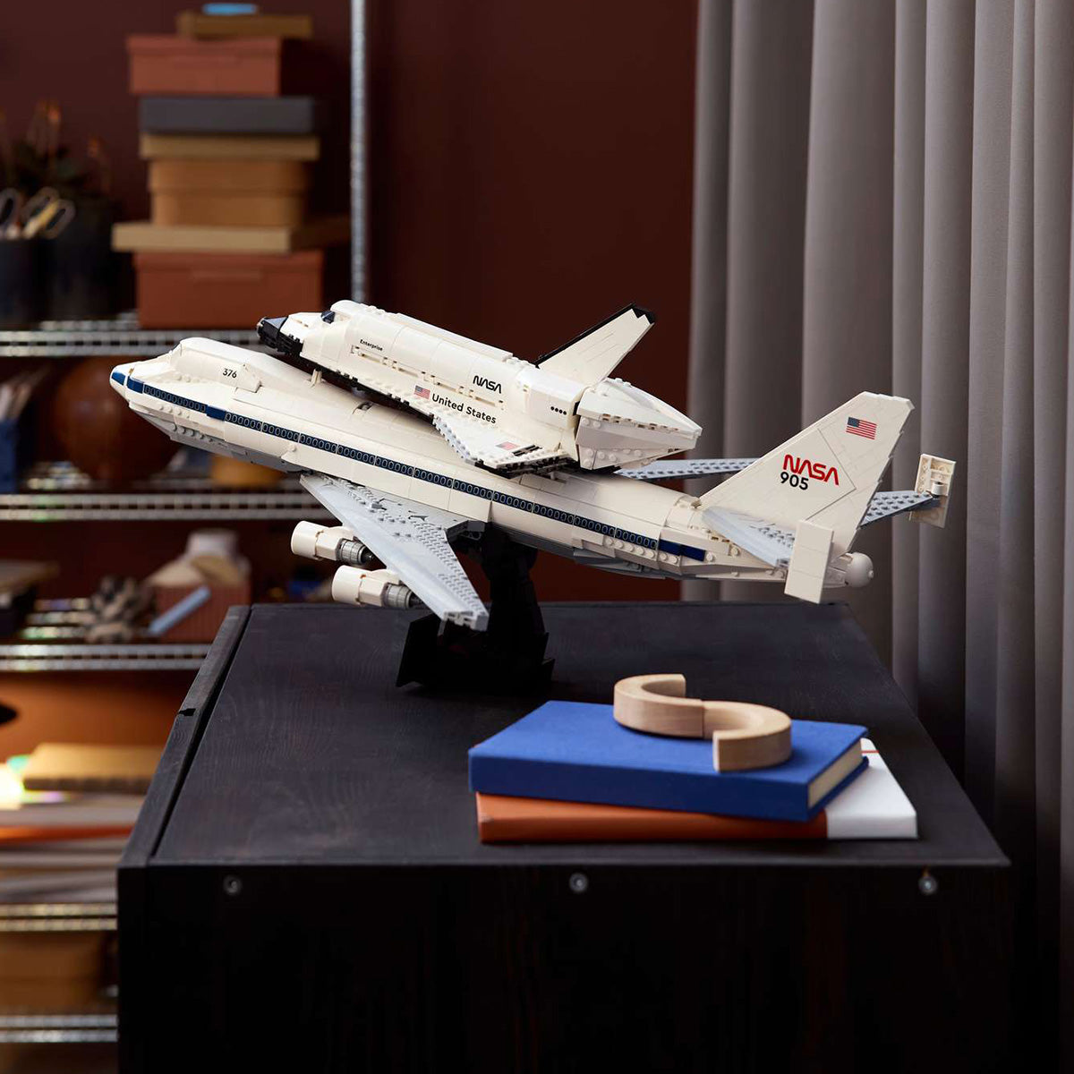 A detailed LEGO® Shuttle Carrier Aircraft model by BOEING, showcasing the NASA Space Shuttle Enterprise atop a Boeing 747, is displayed on a black table near books and decor, with shelves and gray curtains in the background.