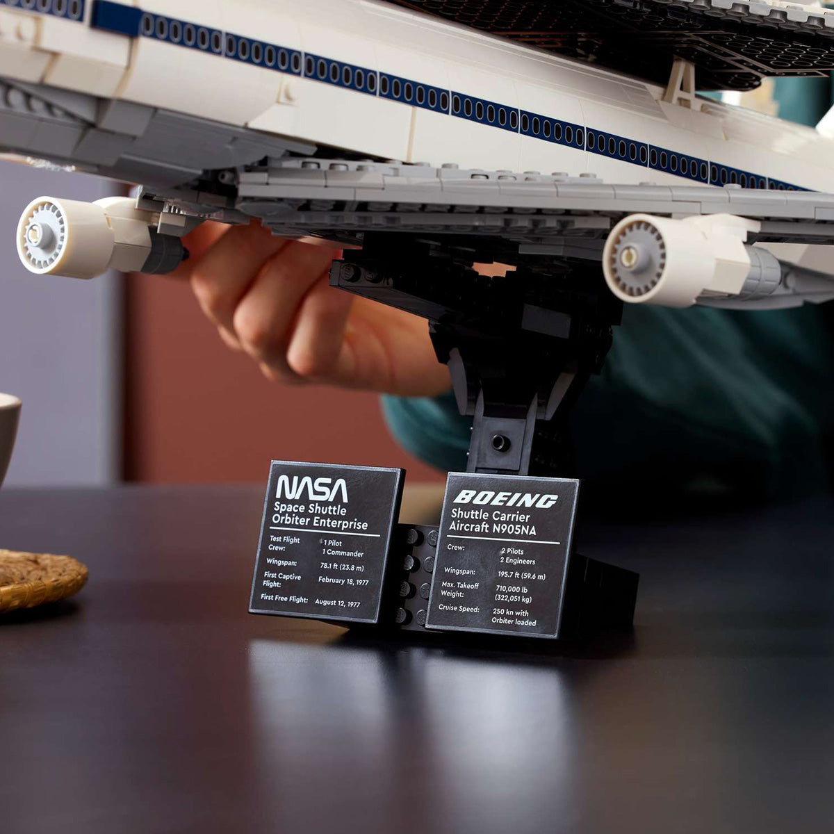 A close-up of a LEGO model featuring the BOEING LEGO® Shuttle Carrier Aircraft, with the NASA Space Shuttle Enterprise mounted on top and displayed on a black stand, accompanied by two info plaques for both the NASA Space Shuttle Enterprise and the BOEING LEGO® Shuttle Carrier Aircraft—a detailed depiction of the Boeing 747 that famously transported the shuttle.