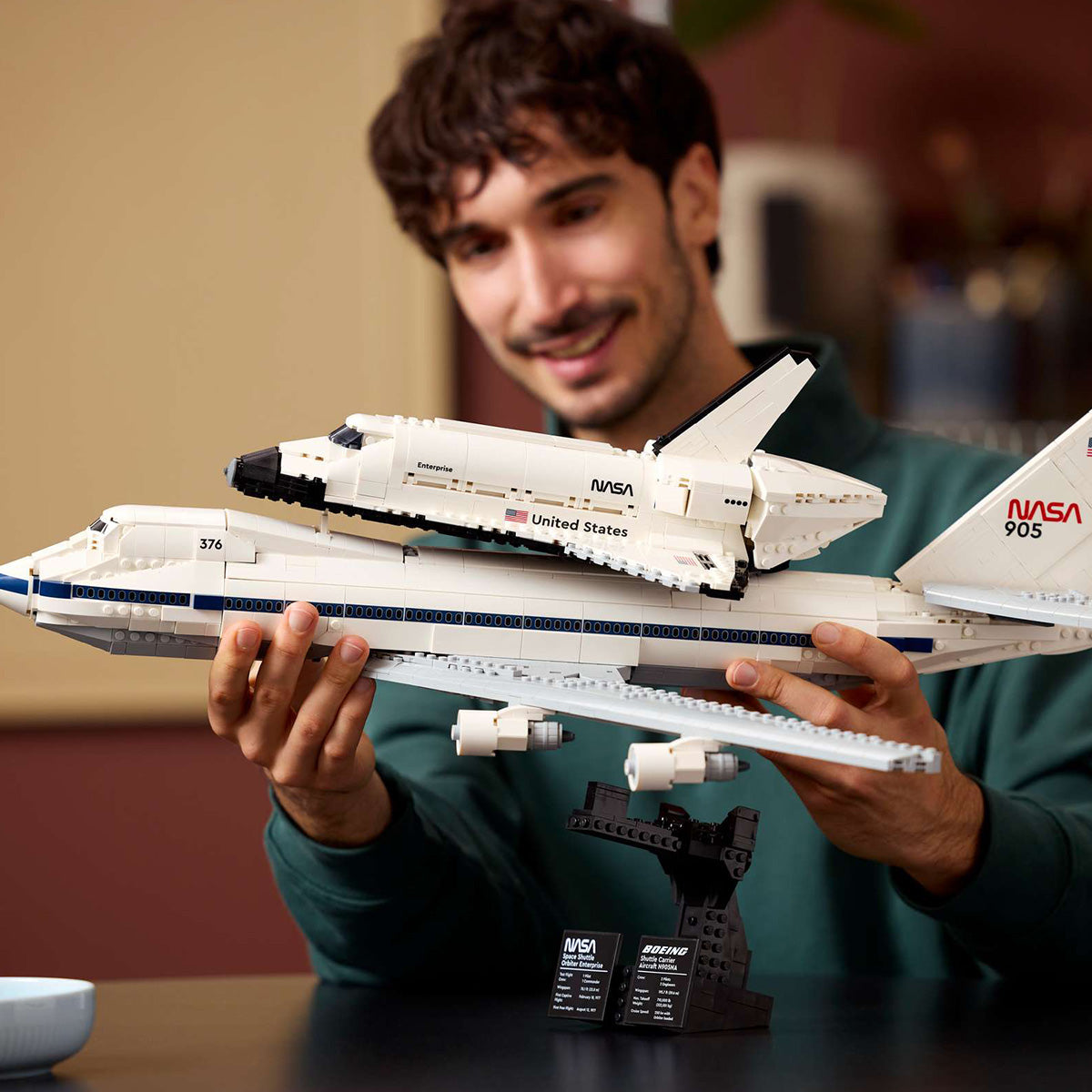 A smiling person holds the BOEING LEGO® Shuttle Carrier Aircraft, which features the NASA Space Shuttle Enterprise mounted atop a Boeing 747, with a display stand visible beneath the model.