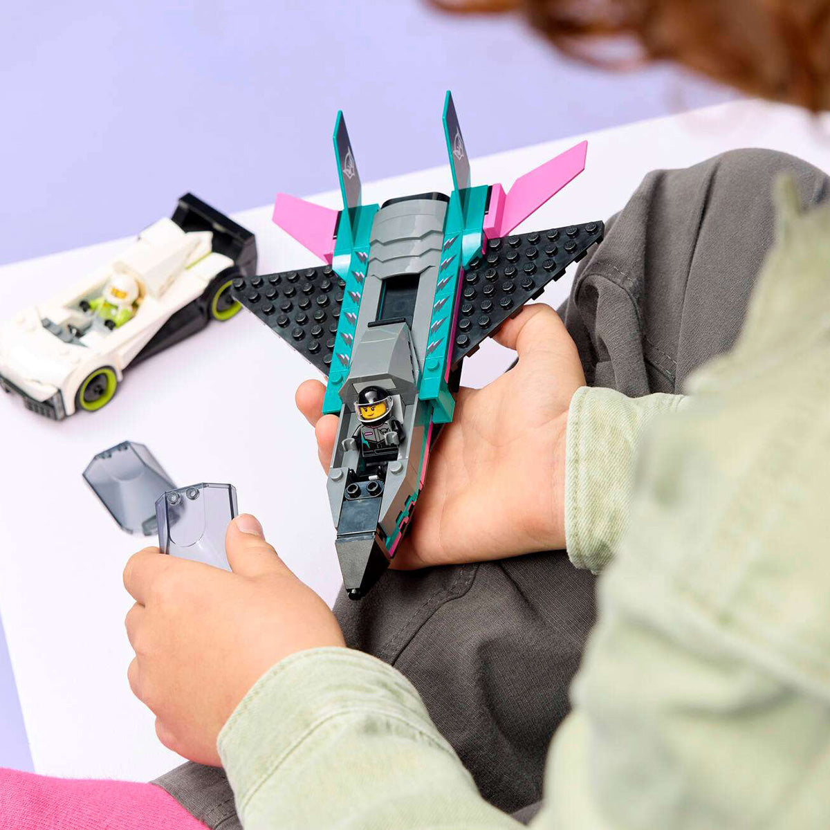 A child holds a BOEING LEGO® Jet vs. Car supersonic jet, featuring vibrant teal and pink accents, and carefully places a canopy over the minifigure seated in the cockpit. Next to them, a sleek white supercar stands poised on a lavender and white surface, ready for racing thrills with the exciting BOEING LEGO® Jet vs. Car playset.
