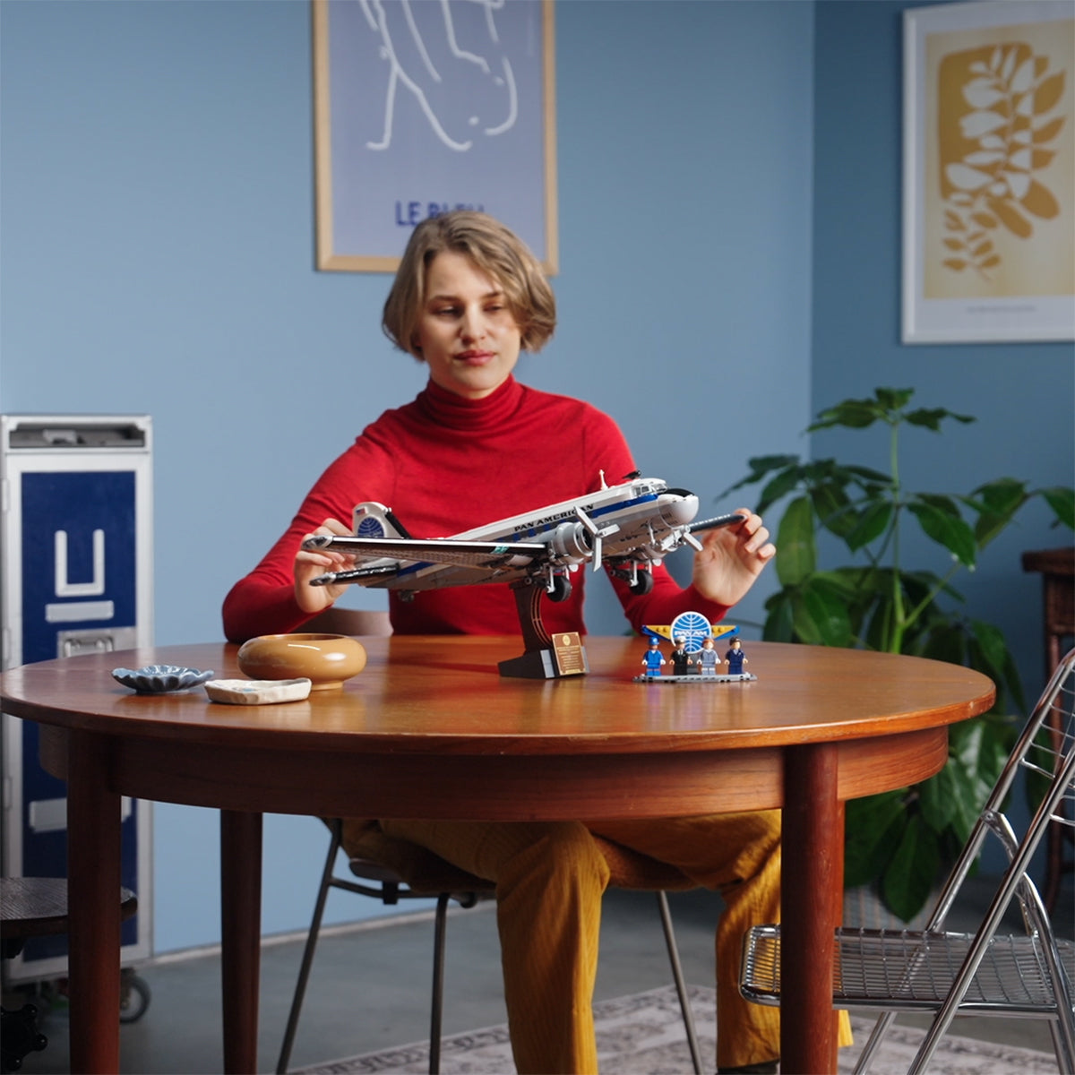 A woman in a red turtleneck assembles the LEGO® Boeing Douglas DC-3 PAN AM Airliner by BOEING at a round wooden table, which also displays a small model, pastry, and tray. Blue walls, framed art, and a green plant are visible behind her.