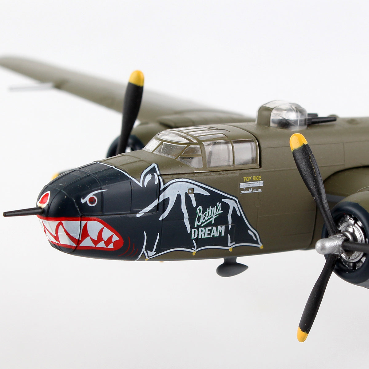 Close-up of DARON's Boeing B-25J Mitchell "Betty's Dream" diecast model, featuring an olive green finish, yellow-tipped propellers, shark mouth nose art, and a white pin-up—an essential collectible for aviation enthusiasts.