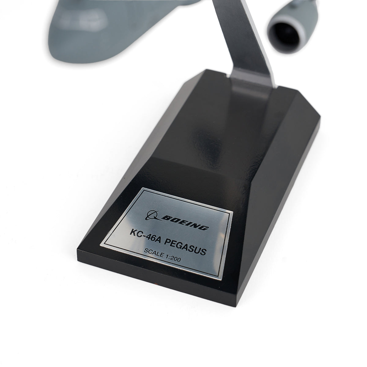 A close-up of a DARON Boeing KC-46A Pegasus 1:200 Model display stand with a metal plaque—ideal for collectors. Part of the model airplane is visible in the background.