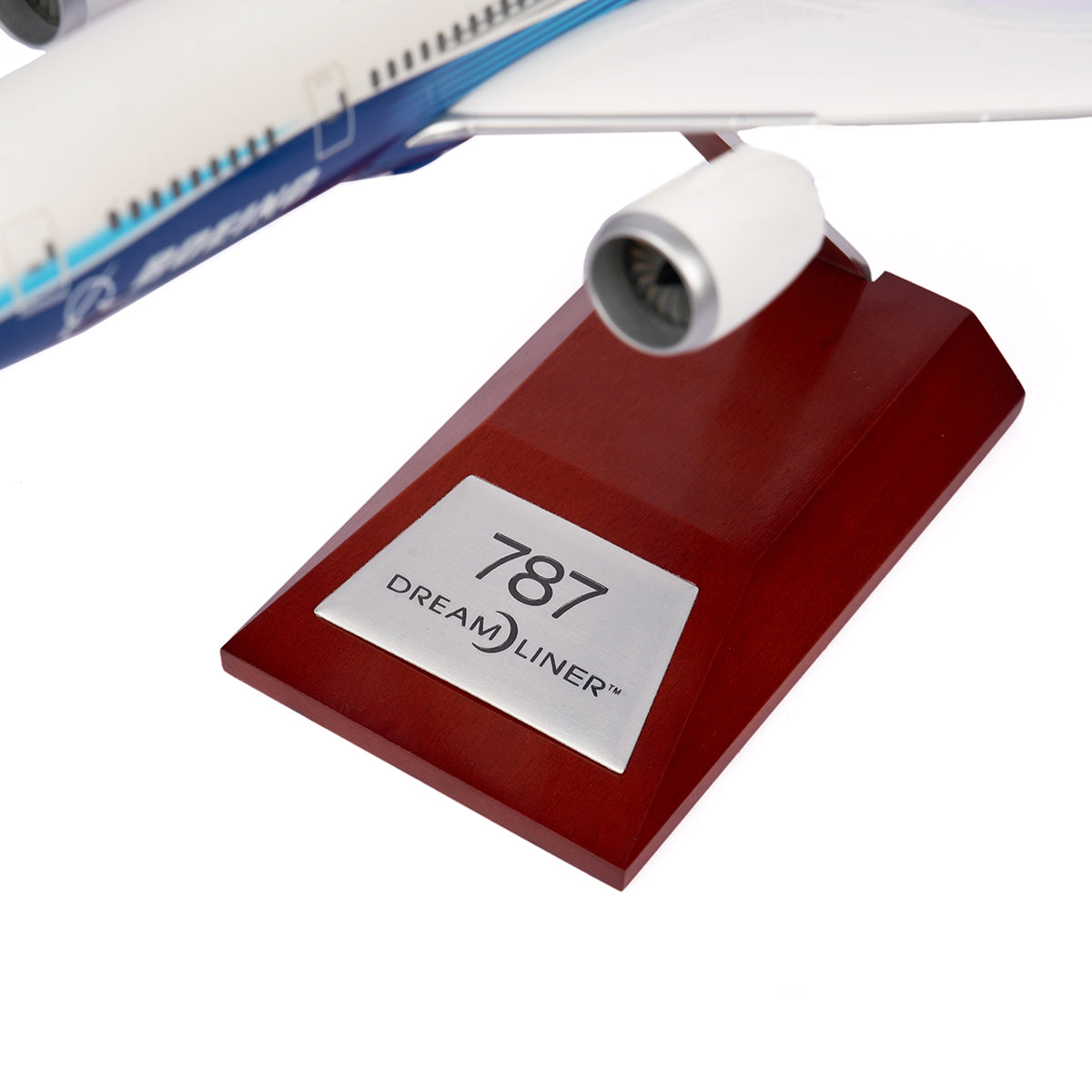 The Boeing Unified 787-8 Dreamliner 1:200 Model by The Boeing Store features a wooden base with a metal plaque engraved 787 Dreamliner. Part of the models wing and engine, symbolizing modern aviation, is visible above the base.