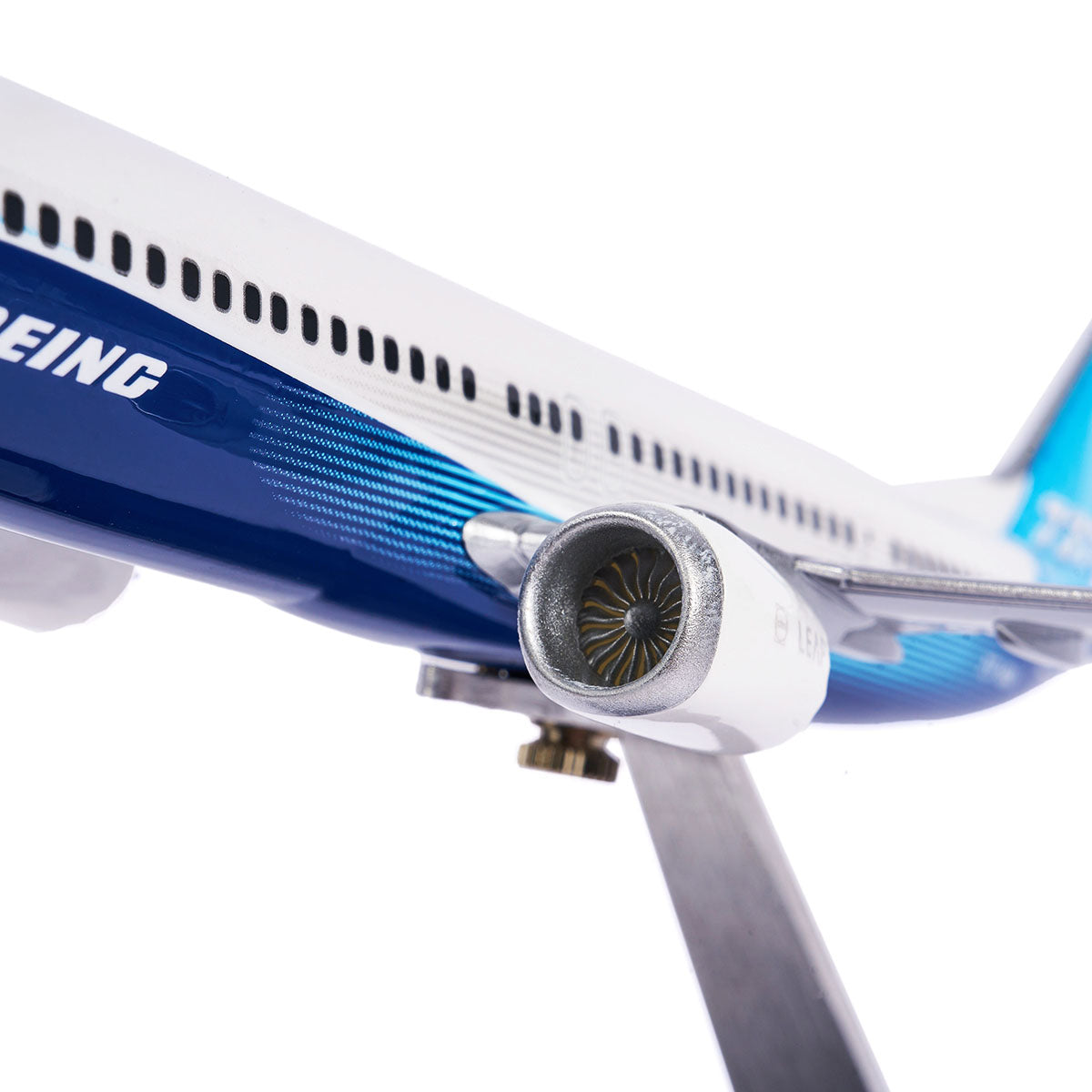 Close-up of The Boeing Stores Boeing Unified 737 MAX 10 1:144 Model airplane with a jet engine on a stand. Its blue and white fuselage displays the Boeing Sky design, highlighting advanced technology winglets against a plain white background.