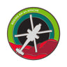 The Boeing AH-64 Apache Skyward Magnet, by Boeing, is a circular patch with a helicopter silhouette and Boeing AH-64 Apache at the top. It features green and red borders with abstract red shapes at the bottom, showcasing an enamel Skyward design aesthetic.