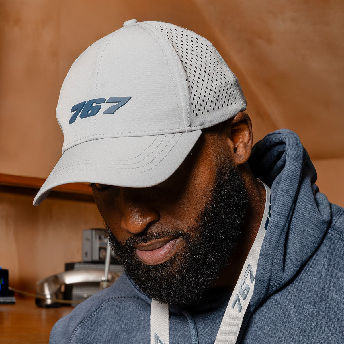 A man wearing a light gray BOEING 767 Series Hat with a matching lanyard looks down. He has a beard and is dressed in a blue hoodie, with a desk and phone blurred in the background.
