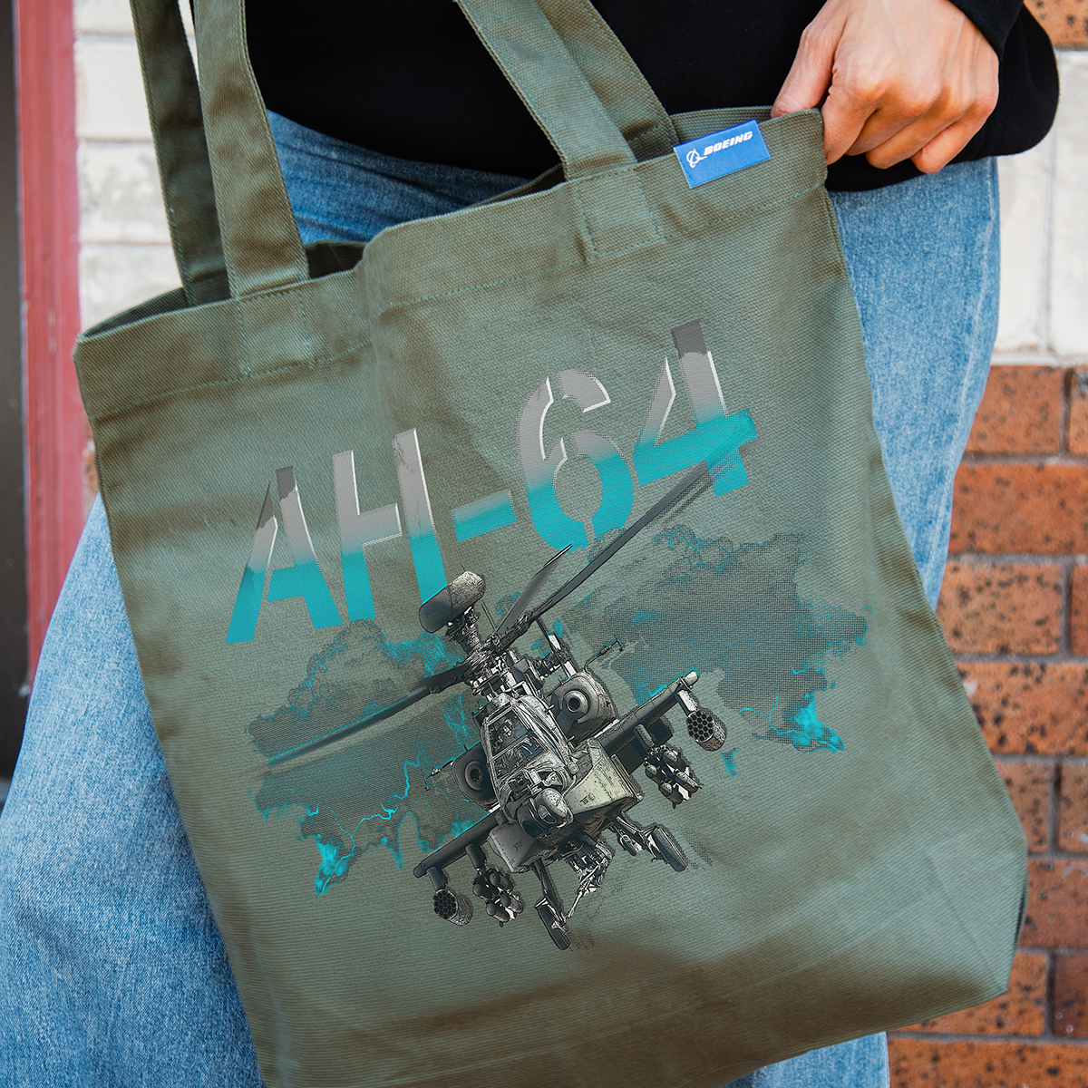 A person holds the Boeing AH-64 Apache Graphic Tote, featuring a helicopter illustration, bold "AH-64" text above, and a BOEING logo tag—ideal for aviation fans and BOEING brand enthusiasts.