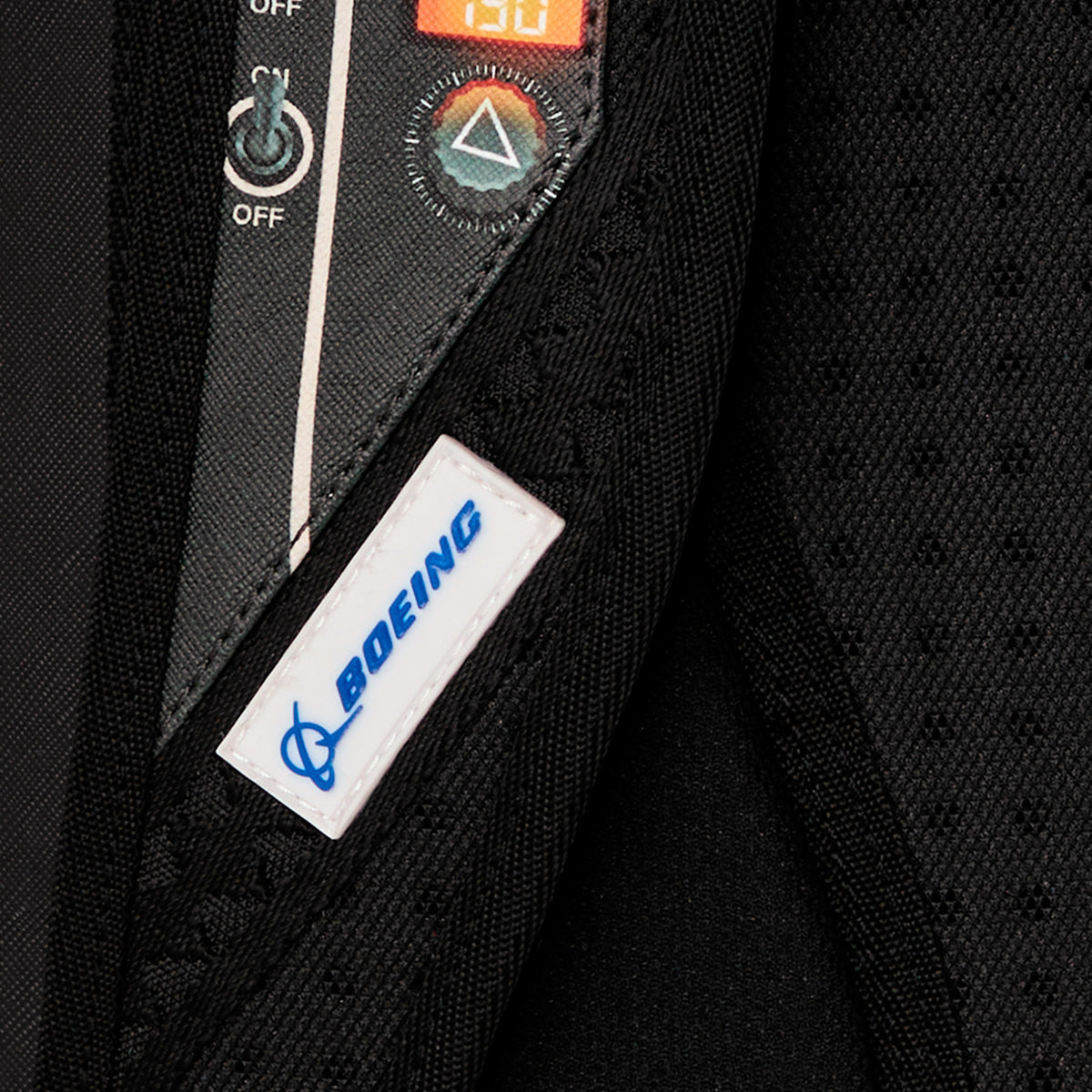 Close-up of a black fabric item, likely the Sprayground Boeing F/A-18 Super Hornet DLXSR Backpack by BOEING, featuring a white "Boeing" label and a control panel with buttons, a triangle symbol, and an LED display showing “130.” This sleek backpack design may also include practical features such as a laptop compartment, making it ideal for use as carry-on luggage.