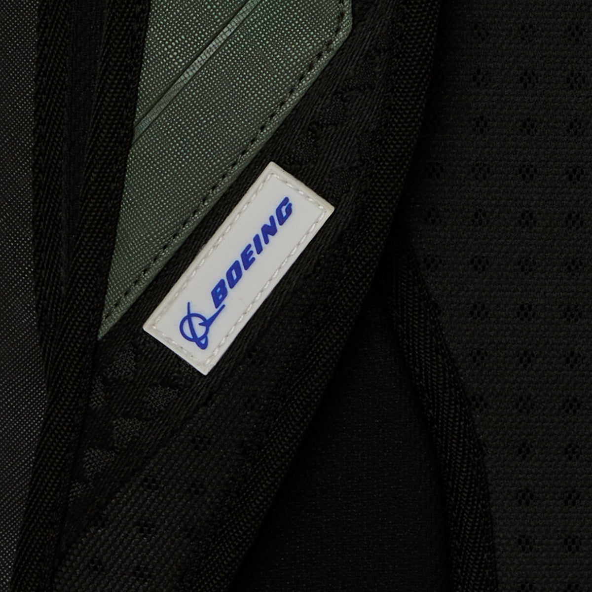 Close-up of a fabric tag featuring the blue BOEING logo and text, stitched onto the black textured surface of the Sprayground Boeing AH-64 Apache Sharkmouth DLXSV Backpack, with hints of green and gray in the background that evoke its aviation heritage and tactical styling.