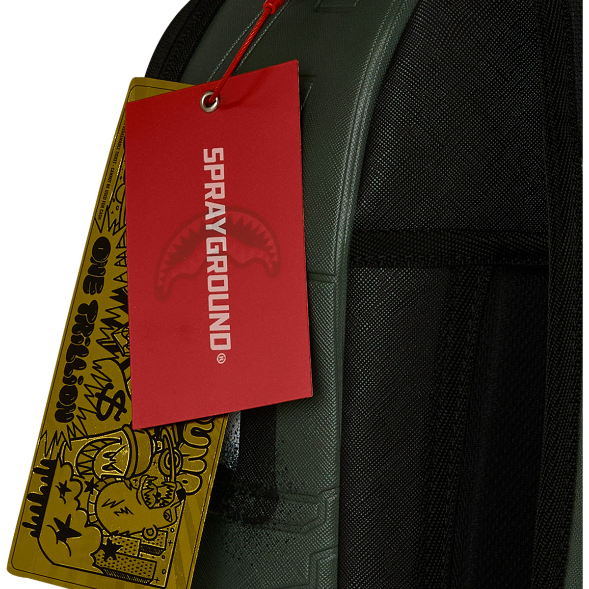 A close-up of the Sprayground Boeing AH-64 Apache Sharkmouth DLXSV Backpack by BOEING, featuring two tags: a red Sprayground tag and a gold tag with cartoonish graffiti-style art displaying the words ONE TRILLION, showcasing the brand's bold design and subtle tactical styling.