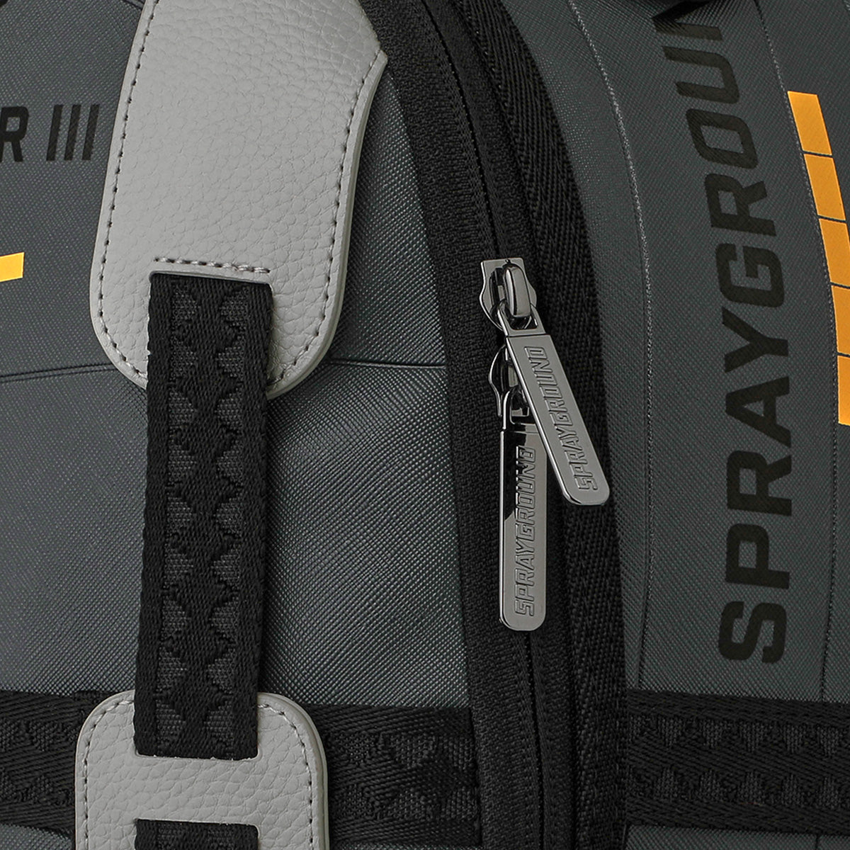 Close-up of a gray Sprayground Boeing C-17 Globemaster DLXSV Backpack, featuring zipper pulls with the BOEING brand name and a portion of the vertical SPRAYGROUND logo on the fabric. Black and textured gray straps accentuate the military-inspired design of this backpack.