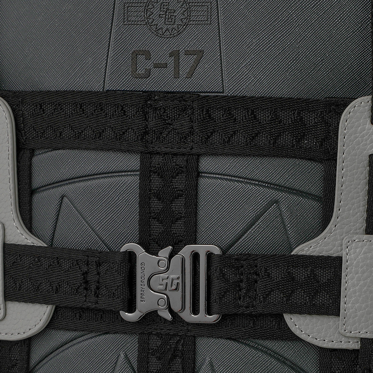 Close-up of a gray fabric with “C-17” text, black woven straps, a metallic SG buckle, and textured light gray leather accents, displaying the detailed craftsmanship of the Sprayground Boeing C-17 Globemaster DLXSV Backpack from BOEING—a military-inspired piece.