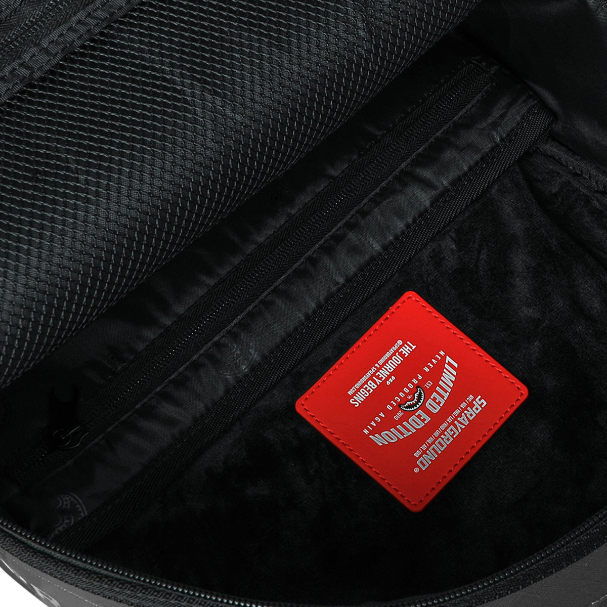The Sprayground Boeing C-17 Globemaster DLXSV Backpack by BOEING, shown open in black, features a zippered pocket, mesh compartment, and a red rectangular label with white text and graphics sewn onto the interior fabric, emphasizing its military-inspired design.