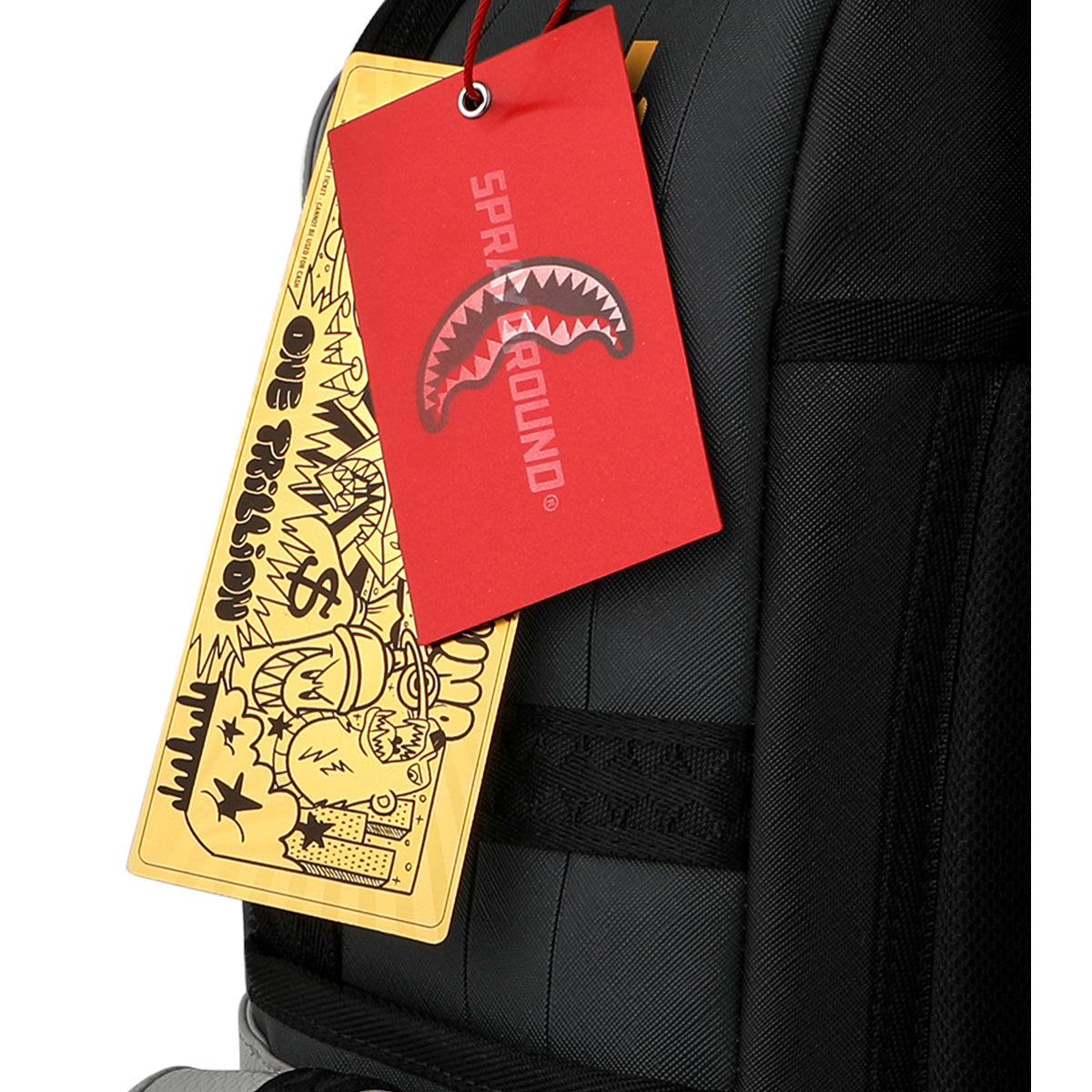 Close-up of the Sprayground Boeing C-17 Globemaster DLXSV Backpack by BOEING, a black, military-inspired design featuring two tags: one red SPRAYGROUND tag with a shark mouth graphic and one yellow comic-themed tag with illustrations and the text "ONE TRILLION.