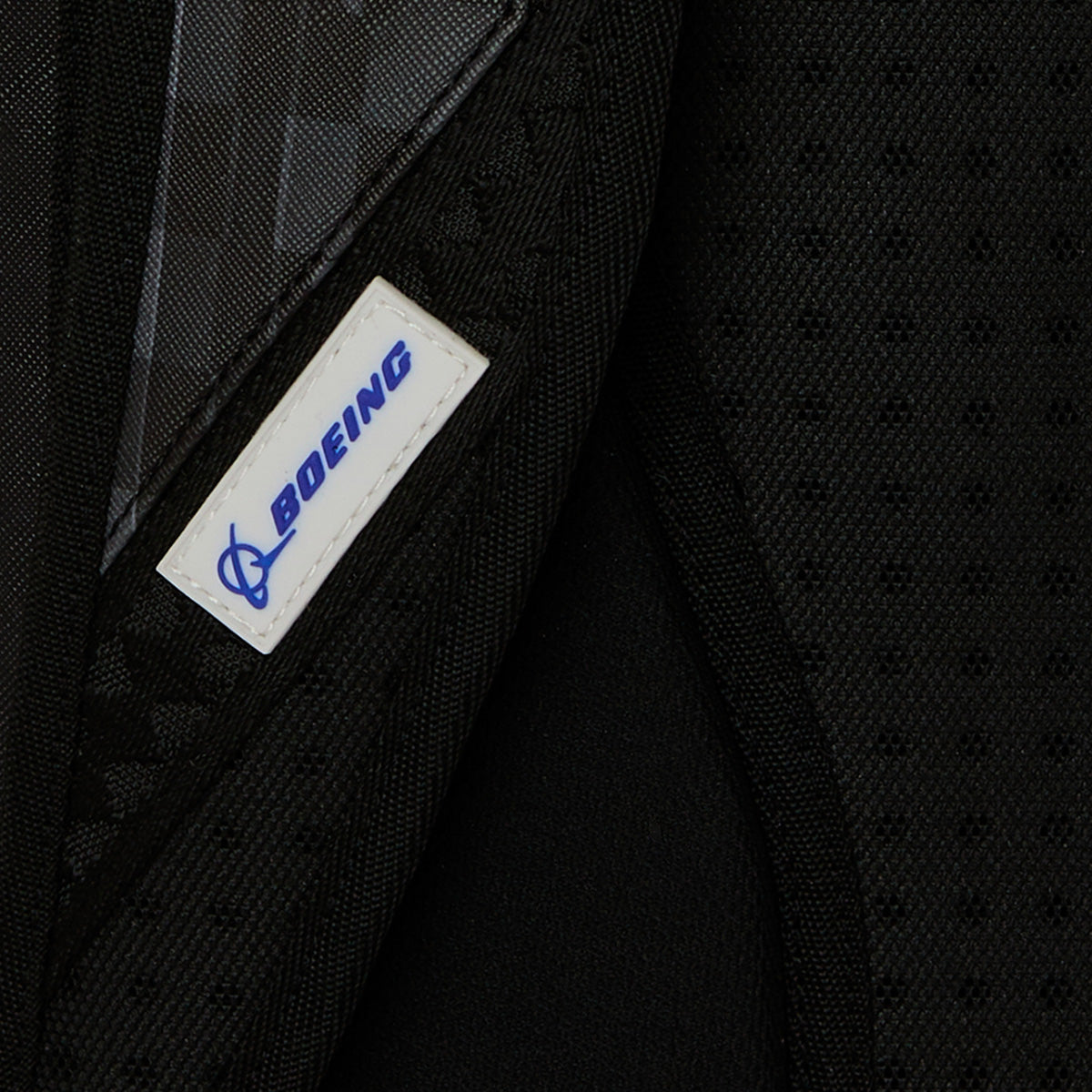 Close-up of a black fabric from the BOEING Sprayground P-51 Mustang Sharkmouth DLXSV Backpack, featuring a white and blue Boeing logo label stitched onto it. The fabric appears padded and textured, reflecting the specialized design of this aviation-inspired backpack.