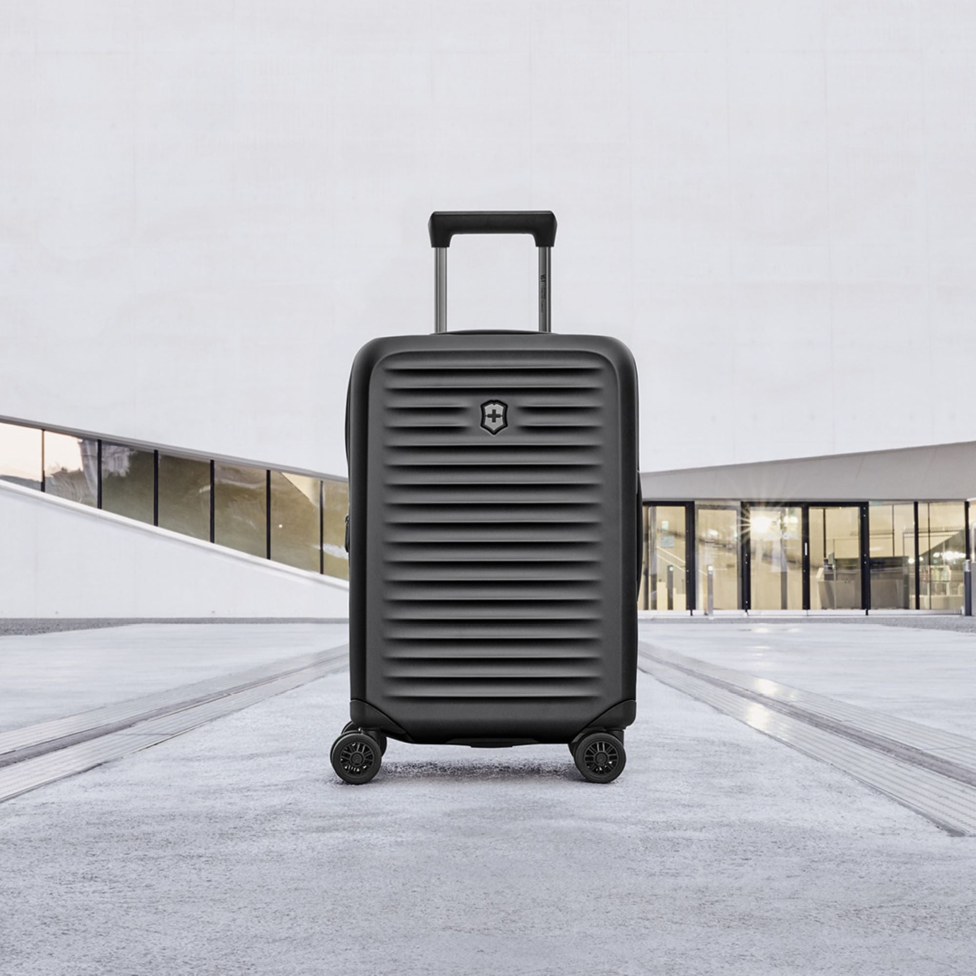 The Victorinox Airox Advanced Frequent Flyer Carry-on in black, featuring dual caster wheels and a ribbed modern design, stands upright on a concrete surface. Its lightweight build is ideal for travel. The background of modern architecture with large windows and a smooth white exterior adds sophistication.
