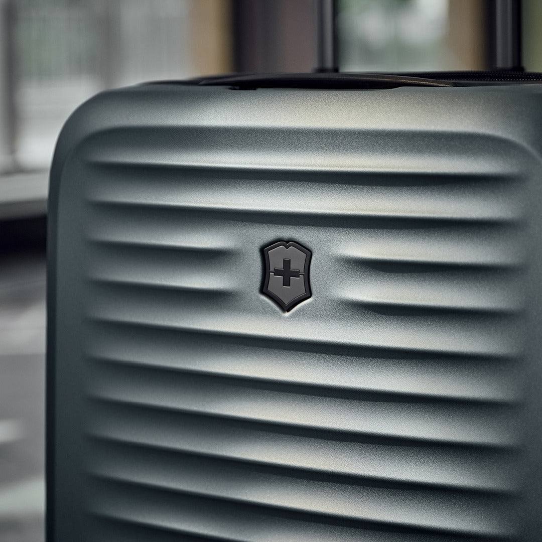 Close-up of textured, metallic Victorinox Airox Advanced Frequent Flyer Carry-on - Storm, featuring a small central emblem with a cross. This VICTORINOX model has dual caster wheels for smooth travel. The blurred indoor background suggests refined design elements.