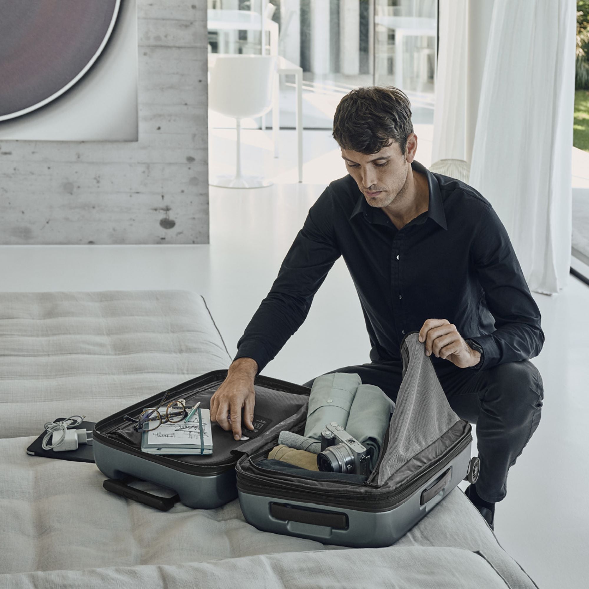 A person in a black shirt is packing their VICTORINOX Airox Advanced Frequent Flyer Carry-on - Storm, featuring a dual caster wheel system, on light-colored sheets. Clothes and a camera are packed inside, with glasses and a watch nearby, complementing the rooms modern minimalist design.