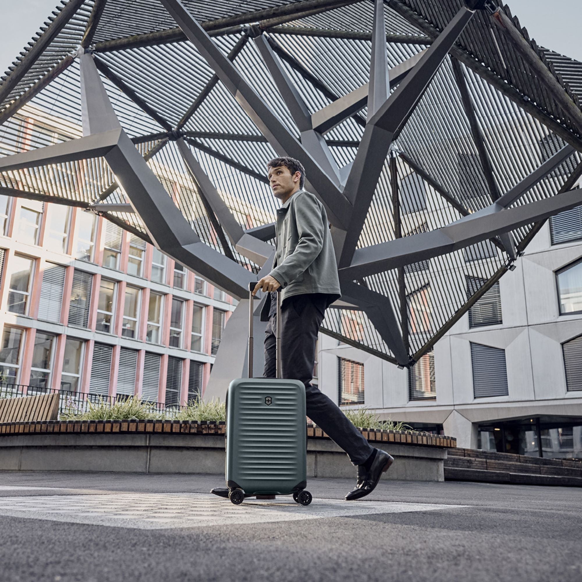 Dressed in a green jacket and black pants, a person walks outside with the Victorinox Airox Advanced Frequent Flyer Carry-on - Storm. They pass under a large geometric metal sculpture, set against modern buildings.