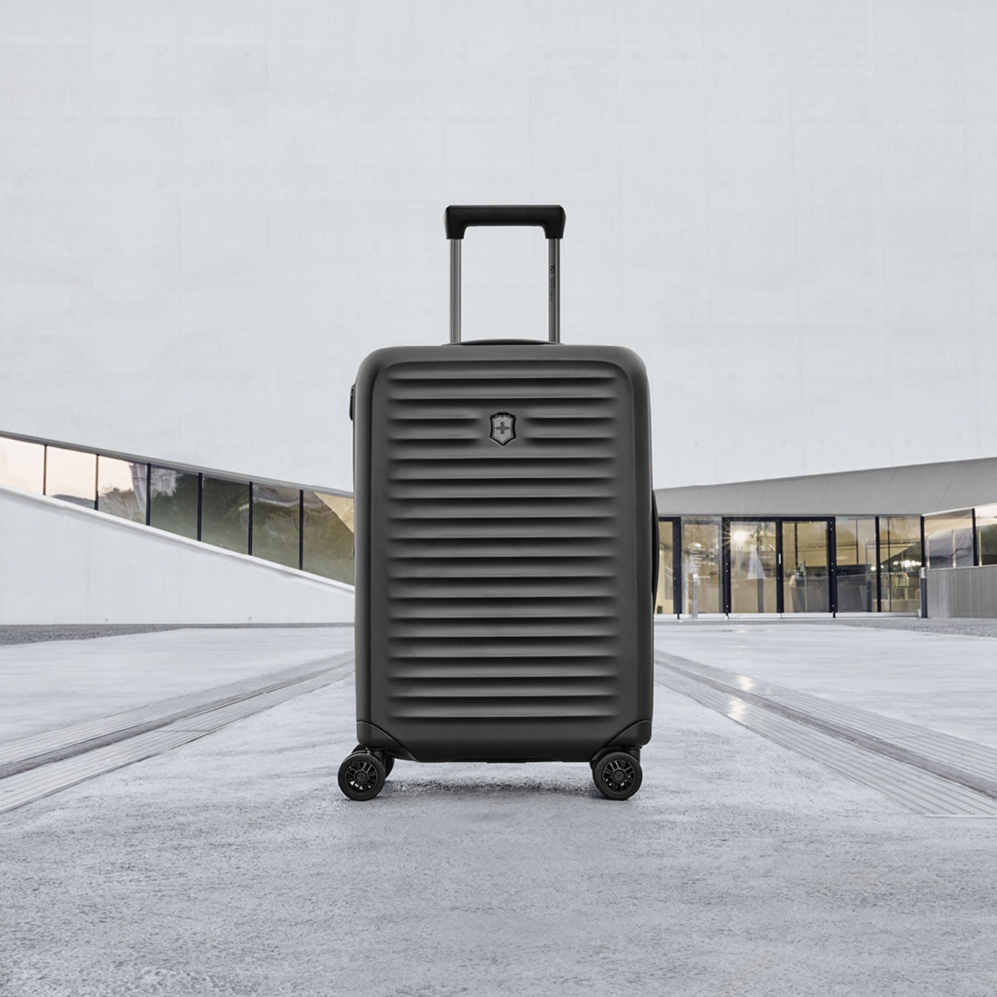 The Victorinox Airox Advanced Frequent Flyer Carry-on Plus - Black, with a ribbed design and four wheels, stands on concrete. Designed for frequent flyers, its lightweight build promises travel ease. A modern building with large glass windows is the backdrop as its telescopic handle is extended.