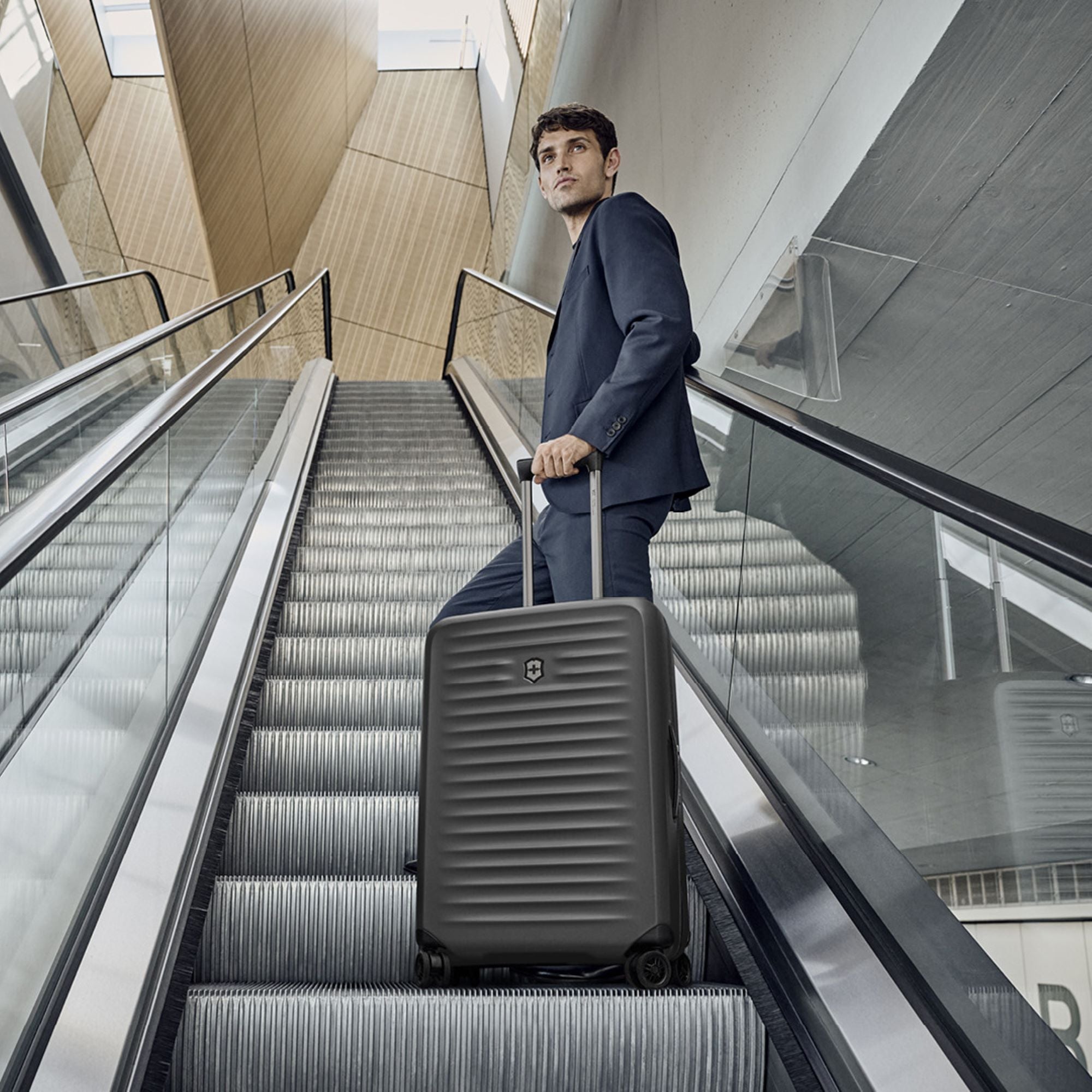 A man in a blue suit, carrying the Victorinox Airox Advanced Medium - Black luggage by VICTORINOX with its anti-microbial lining, stands on an escalator in a modern building. He gazes up as natural light highlights the wood and concrete architectural design.