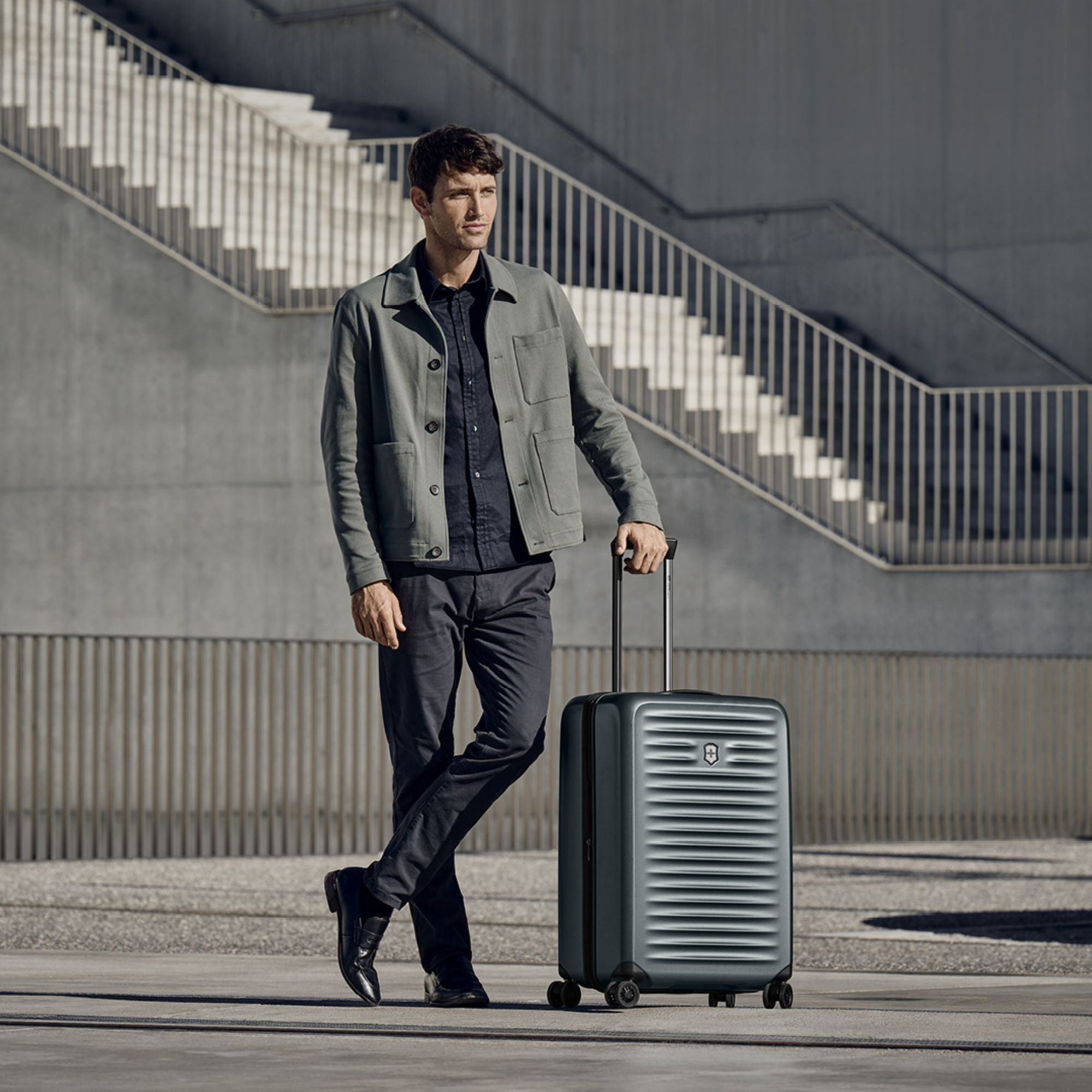 A man in a gray jacket and dark pants walks with his Victorinox Airox Advanced Medium - Storm luggage by VICTORINOX in front of a modern, industrial-style building featuring stairs and railings.