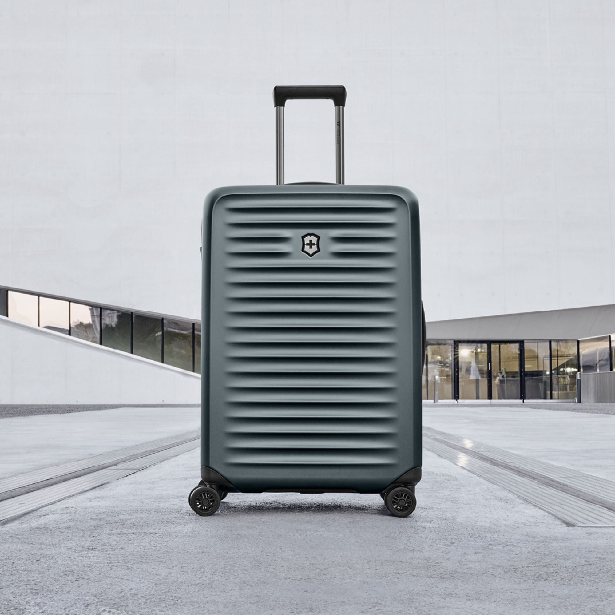 The Victorinox Airox Advanced Medium - Storm, by VICTORINOX, is a dark gray, ribbed hard-shell suitcase with anti-microbial lining. Featuring a telescopic handle and four wheels, it stands upright on concrete. The modern buildings large windows and curved facade complement this lightweight luggage.