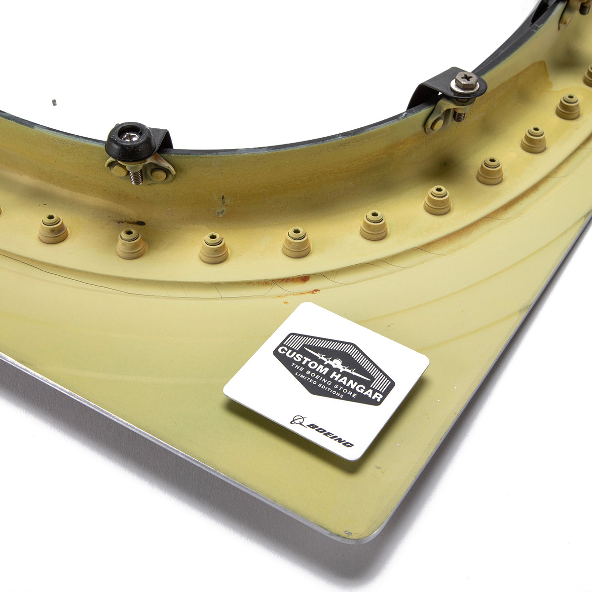 A close-up of a curved, yellow-green Boeing 777 Natural Finish Window with bolts and fasteners, featuring a square "Custom Hangar by Boeing" sticker—an authentic aviation artifact.