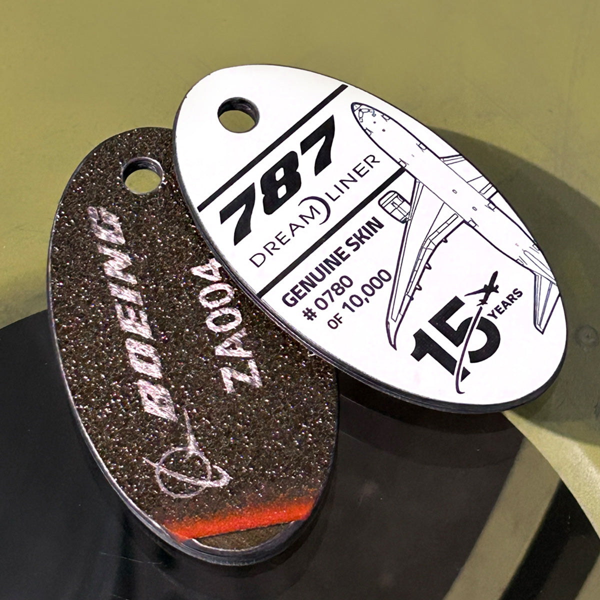 Two BOEING PlaneTags from the Boeing 787 Series: one is inscribed with ZA004, while the other displays “Boeing 787 Dreamliner,” “Genuine Skin #0780 of 1000,” and “15 Years.” Both tags are crafted from authentic carbon-fiber composite material sourced from the Dreamliner and feature holes designed for keychain attachment.