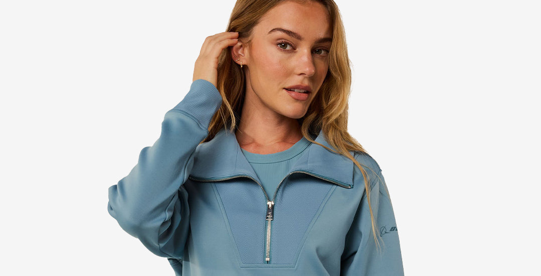 A female model wearing a light blue zip-up sweatshirt, with her hand touching her hair, against a clean white background.