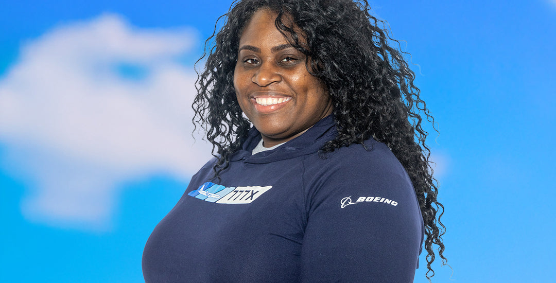 A person wearing a navy blue Boeing Velocity sweatshirt with the Boeing logo on the sleeve, standing against a blue sky with clouds.