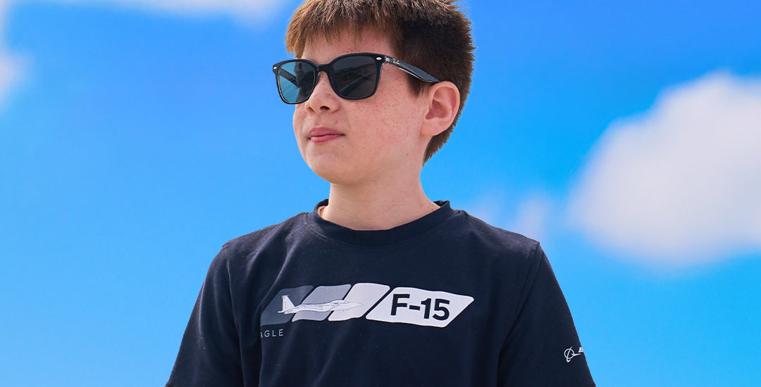 A child wearing sunglasses and a black Boeing Velocity t-shirt featuring an F-15 design, standing outdoors with a blue sky in the background.
