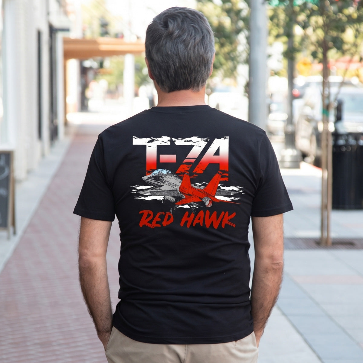 A man on a city sidewalk wears the Boeing T-7A Red Hawk Unisex Graphic T-Shirt by BOEING, featuring a red and gray airplane, clouds, and “T-7A RED HAWK” text on the back—a great choice for aviation fans.