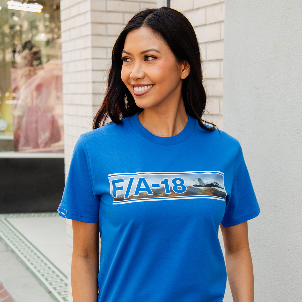 A woman with long dark hair smiles outdoors by a light brick wall, wearing the BOEING F/A-18 Super Hornet Unisex Graphic T-Shirt featuring a jet graphic.