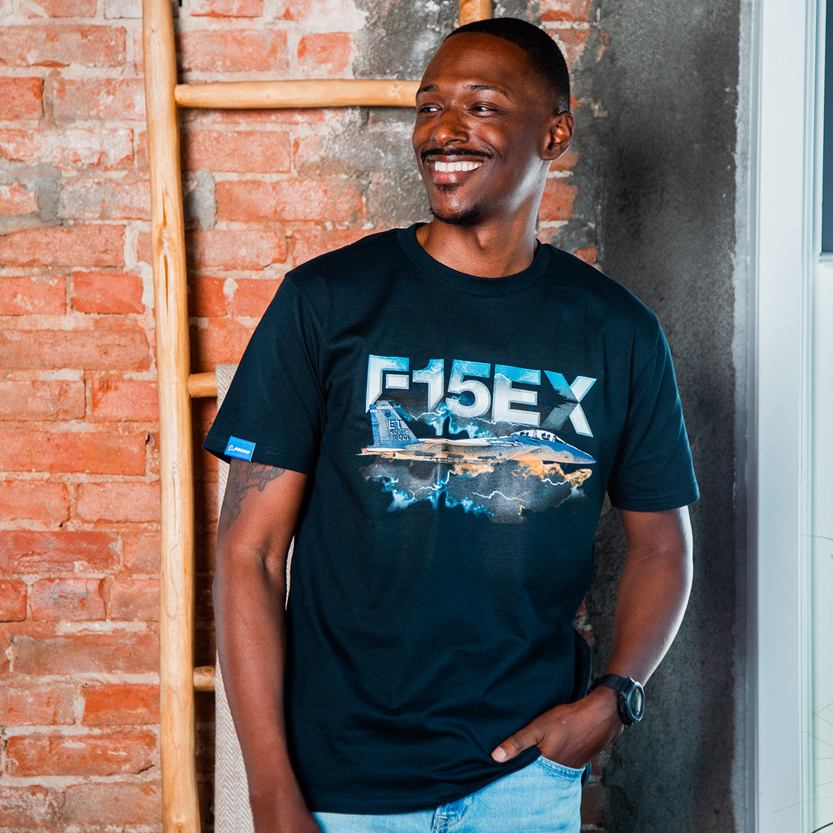 A smiling man stands by a brick wall and wooden ladder, wearing a BOEING F-15EX Eagle II Unisex Graphic T-Shirt with a fighter jet design—an ideal choice for aviation enthusiasts.