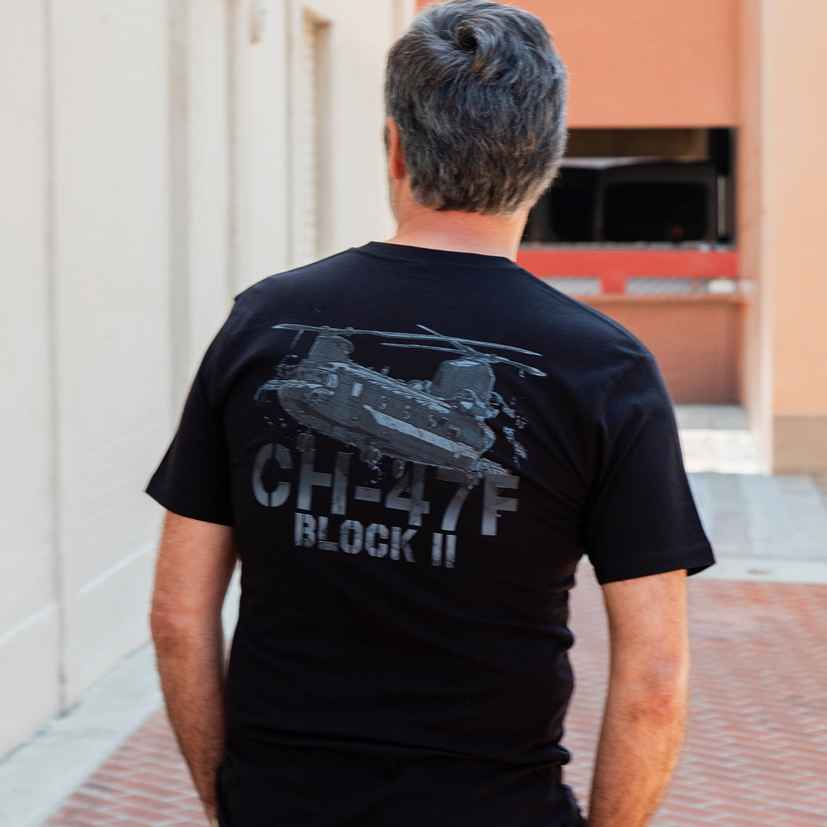 A man with gray hair stands in an alleyway, facing away. He wears a Boeing CH-47F Block II Unisex Graphic T-Shirt—a great pick for aviation fans and supporters of the BOEING brand.