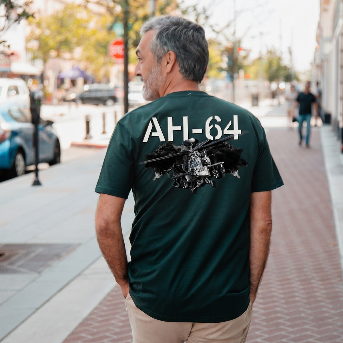 A man with gray hair stands on a city sidewalk wearing the BOEING AH-64 Apache Unisex Graphic T-Shirt, featuring a military helicopter design—ideal for aviation fans and supporters of Boeing.