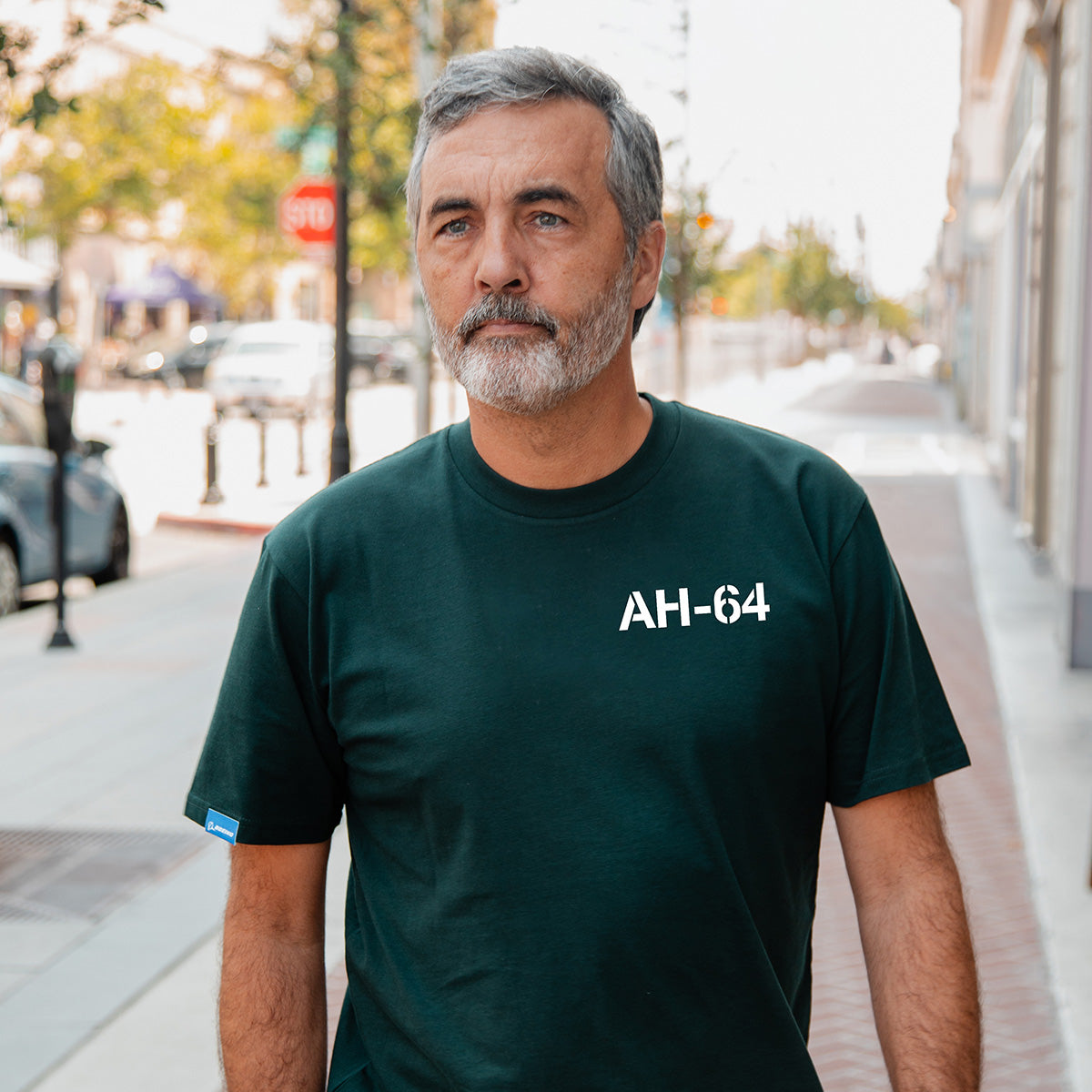 A man with gray hair and a beard stands on a city sidewalk wearing the Boeing AH-64 Apache Unisex Graphic T-Shirt by BOEING—ideal for aviation fans. Trees and parked cars line the street in the background.