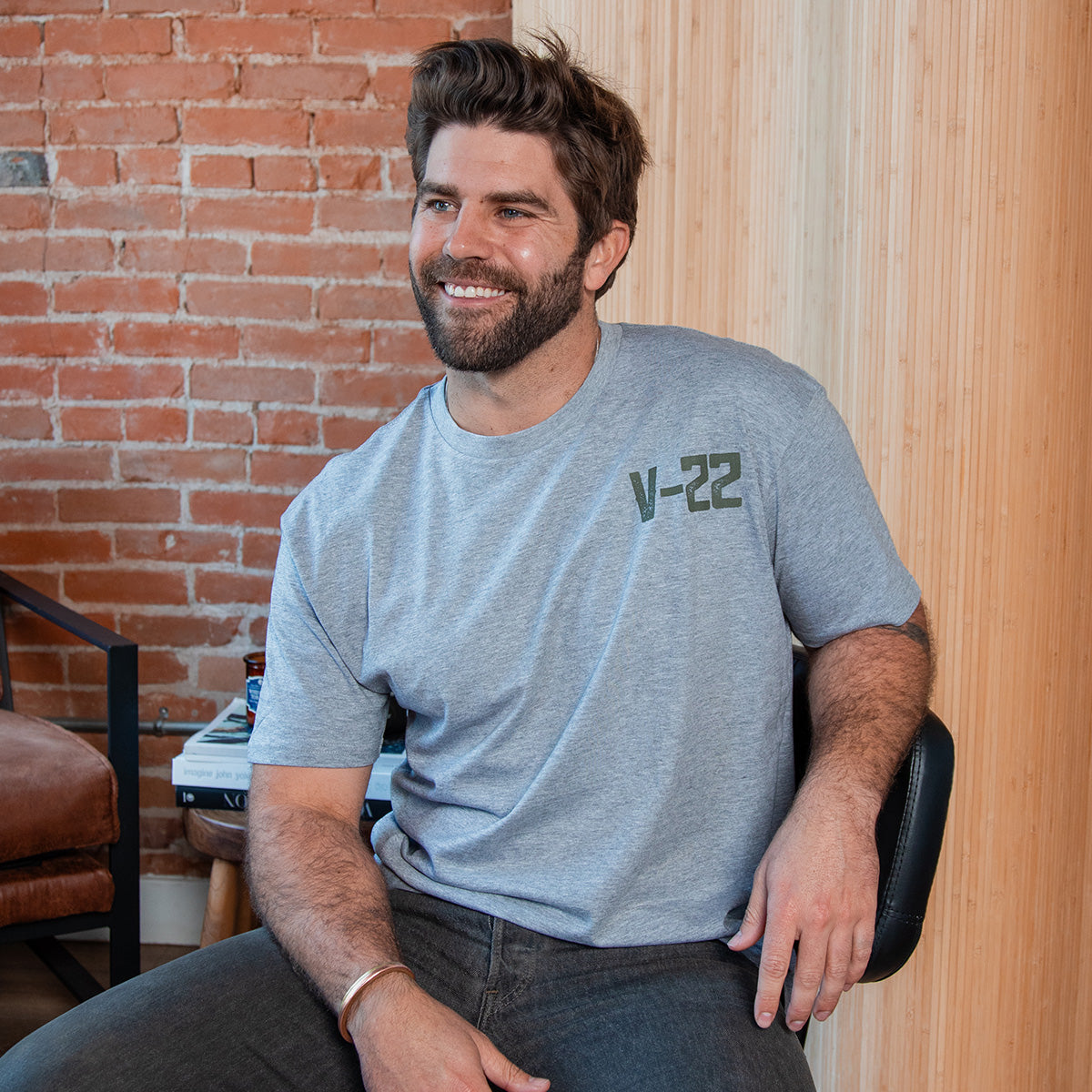 A smiling man with brown hair and a beard sits on a chair in a modern room with a brick wall, wearing the BOEING V-22 Osprey Unisex Graphic T-Shirt and dark jeans.