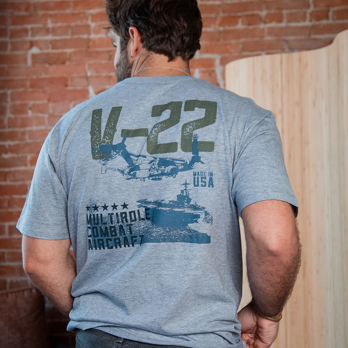 A man faces away in a light gray Boeing V-22 Osprey Unisex Graphic T-Shirt by BOEING, featuring an aircraft graphic, "MULTIROLE COMBAT AIRCRAFT," Boeing carrier, five stars, and "MADE IN USA" text against a brick wall backdrop.