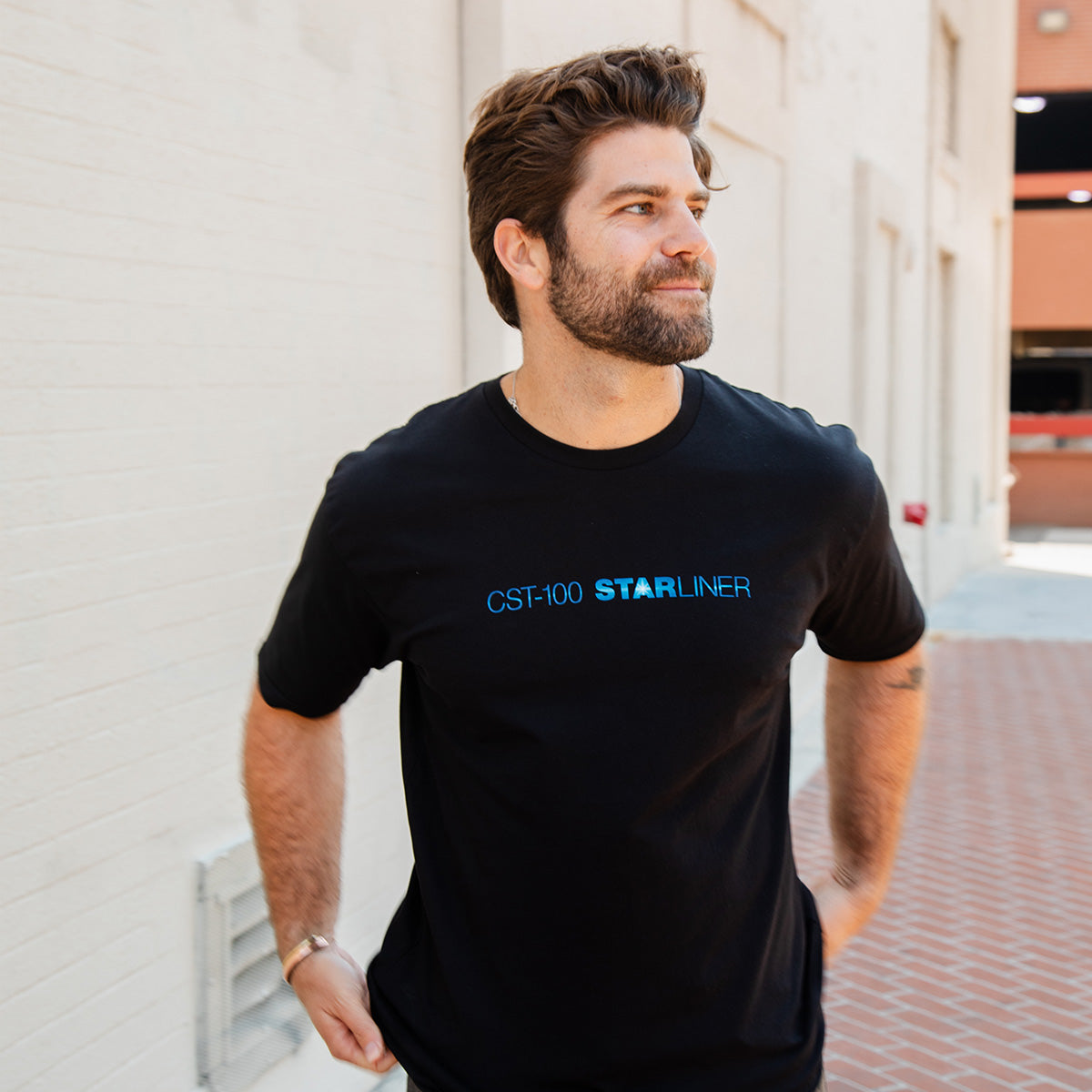 A man with brown hair and a beard walks outside on a brick sidewalk, wearing a black BOEING CST-100 Starliner Unisex Graphic T-Shirt. Behind him is a white brick wall and part of a building.
